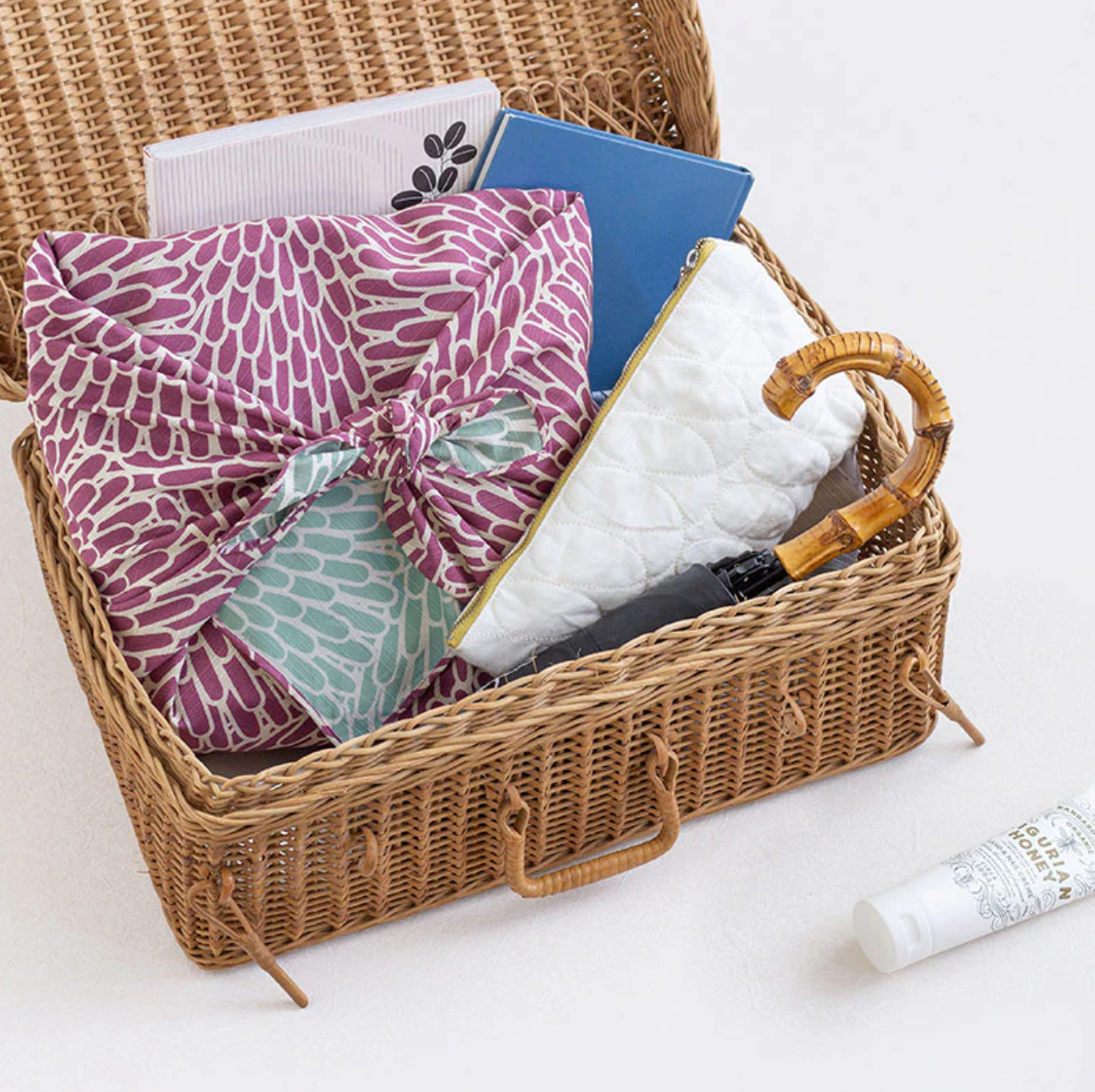 Wicker picnic basket with a purple patterned furoshiki, white pillow, and books on a white background